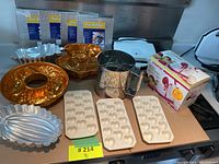 Assorted bakeware items including gold bundt pans, heart-shaped silicone molds, a metal sifter, and four silver pot holders.