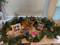 Wide view showing artificial pine garland with string lights, woven light-up star decoration, four colorful spherical ornaments, boxed assorted decorations, and gold bear figurine.