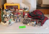 Overall lot showing illuminated ceramic house with snow and figurines, boxed in background, multiple packs of themed figurines and accessories, and poinsettia patterned linens