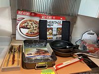Photo showing boxed Revere pizza pans, Heritage Rock frying pans stacked, WearEver roasting pan with rack still in plastic, and carving tool set in box with wooden handles.