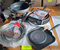 Top view of the pan and skillet set on table showing a mix of black pans, a boxed Gotham Steel pan, a round Rival pan still in plastic wrap with spatula, and a square grill skillet with wooden handle.