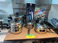 Photo of stainless steel cookware set including multiple pots with glass lids, a small metal pan, boxed pepper grinder and box labeled Grill.