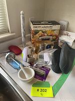 Photo showing boxed Equity Sandwichcrafter Snack'n Sandwich Maker and assorted kitchenware including measuring cups, oven mitts, pizza cutter, and electric mixer on a table near a window.