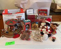 Front view of the Victorian Christmas Carousel in front of its original box with assorted holiday decorations and plush toys arranged around it.