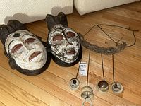 Two carved wooden African style masks placed on wooden floor beside a set of four hanging bells; masks have white paint and red-brown accents; one bell shows damage.