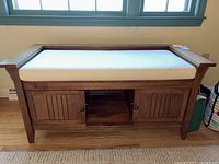 Front view of wooden storage bench with white cushioned seat. Two doors with paneled design and knobs below central open shelf.