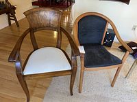 Two wooden armchairs side by side on a hardwood floor and partially on a carpet; one with a carved wooden back and white upholstered seat showing stains, the other with curved wood frame and black upholstered seat and back.