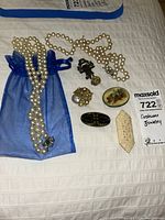 Lot of costume jewelry including two pearl necklaces and six pins displayed on a white surface with a blue mesh pouch.