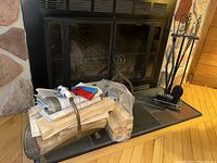 Photo showing the metal fireplace tool stand with poker, broom, shovel, tongs with ceramic handles, and hammered metal log holder filled with firewood pieces and a wrapped newspaper bundle.
