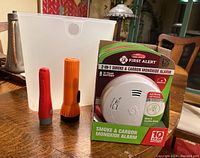 Lot displayed on wooden table showing four First Alert Smoke and Carbon Monoxide Alarms in packaging, two Eveready flashlights, and translucent plastic bin.