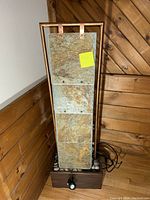Front view of indoor water fountain showing metal frame and four textured stone panels mounted vertically on dark base with decorative stones. Power cord and pump visible at bottom.
