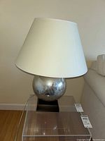 Full view of the lamp showing silver crackled globe base, black wood base, and white shade placed on clear acrylic table.