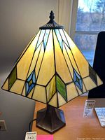 Stained glass table lamp with illuminated shade showing geometric patterns including blue, yellow, green and cream colors on a metal base, placed on a wooden surface by a window.