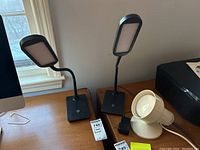 Photo shows two black rectangular LED desk lamps with flexible necks, square bases, and a white clip-on lamp with a round head and mounting clamp on a wooden desk surface near a window.