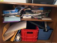 Full view of wooden cabinet shelf filled with various paper supplies including paper reams, photo paper, folders, envelopes, and a red plastic crate with additional papers and supplies beneath.