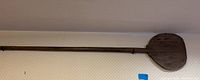 Full view of wooden pizza paddle hanging on wall showing entire length and paddle shape.