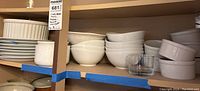 Shelf with stacks of white ceramic bowls and plates along with clear glass bowls, some chipped.