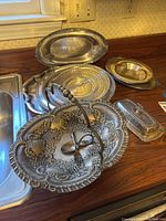 Photo showing various silver plate platters, a decorative serving basket, and a covered butter dish arranged on a kitchen counter.