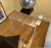Clear acrylic cube table placed on a hardwood floor next to a couch and black and red pillow.