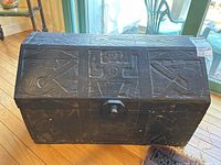 Front view of the dark wood trunk showing carved human figures and geometric patterns with a visible locking mechanism.