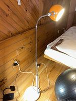 Floor lamp standing next to bed, showing orange lamp shade lit, chrome neck, white base on hardwood floor.