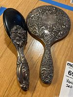 Hand mirror and hairbrush viewed from top showing embossed floral and scroll pattern on silver tone surface and wear on brush edges.