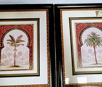 Close-up photo showing the two large framed palm tree prints side by side, highlighting the black frames with gold borders and the artwork within the decorative arch patterns.
