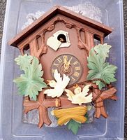 Front of antique wooden cuckoo clock with birds, carved leaves, and chalet style roof top. Shows decorative green leaves, two carved white birds with yellow nest detail, and clock hands showing 12:05.