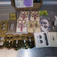 Various electrical outlet and switch wall plate covers laid out showing ceramic floral, brass, white plastic, and painted covers.