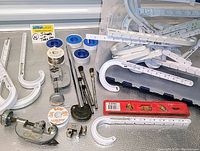 White plastic J-hook support hangers with visible mounting holes, metal pipe clamp, blue tape rolls, magnetic level, wire spool, and brushes laid out on a surface alongside a clear blue tote bin.