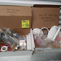 Two open cardboard boxes with various plumbing parts, new pipe accessories, and a set of new white ceramic bathroom holders including towel ring and paper holder.