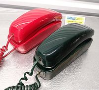 Red and green vintage slim Trimline telephones with coiled cords, shown side-by-side on a metallic surface.