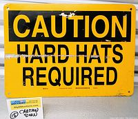 Front view of rectangular yellow metal caution sign with black text 'CAUTION HARD HATS REQUIRED'. Shows wear spots especially near center and corner areas, hole in each corner for mounting.