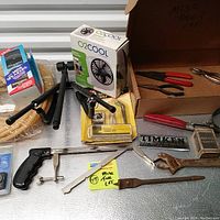 Box lot of miscellaneous hand tools including a portable fan, hand saw, pliers, stapler, wrench, and other items arranged on a surface.