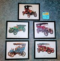Five framed vintage antique car prints arranged on carpet floor showing the full set with black wood frames and visible detailed color illustrations of old cars.