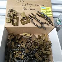 Wide view of box containing numerous metal cabinet hinges and decorative handle pulls on a cardboard base labeled 'Old Vintage Cabinet Hardware'.