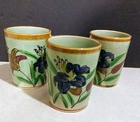 Three vintage Japanese ceramic cups decorated with blue iris flowers and other floral motifs, arranged standing upright to show the painted designs and gold-colored rims.