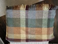 Folded wool blanket with checkered pattern in green, tan, cream, burgundy colors and fringed edges.