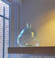 Photo of the blue-tinted clear glass vase on a wooden shelf near a window and wall with tile pattern.