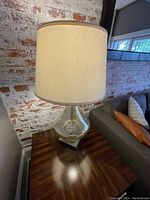 Full view of the lamp on a wooden side table next to a couch, showing the cream fabric shade and glass base.