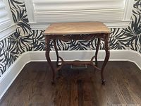Full front view of the antique console table showing carved wooden legs and decorative inlay top surface, placed against a patterned wallpaper background.