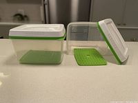 Two clear Rubbermaid FreshWorks rectangular containers with white lids featuring a green vent rim, one lid off showing the clear container and green plastic perforated pad inside.