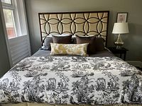View of king-sized bed with black and white Chinoiserie toile duvet cover and cognac/cream upholstered wood framed headboard, showing matching lumbar pillow at front.