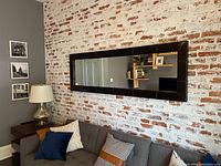Mirror mounted on a distressed brick wall above a grey tufted sofa with various pillows and side table with lamp.