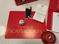 Scattergories game contents including 6 black pencils, answer cards, 20-sided die, timer, and red game folder with logo.