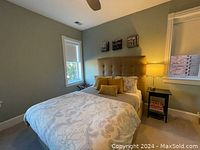 Full view of the queen bed against wall with window, showing tufted beige upholstered headboard with matching fabric base, white patterned bedding and pillows, bedside lamp and nightstand.