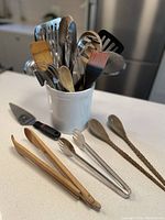 Photo showing the white ceramic crock filled with assorted kitchen utensils including wooden spoons, metal tongs, spatulas, ladles, whisk, meat mallet, and pastry brush.