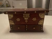 View of the jewelry box front, showing ornate wood finish with brass-tone decorative accents and two small drawers.