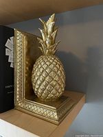 Front and side angle shot of one gold pineapple resin bookend on a shelf with books.