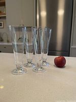 Four clear traditional pilsner glasses shown standing upright next to an apple for size reference on a counter.